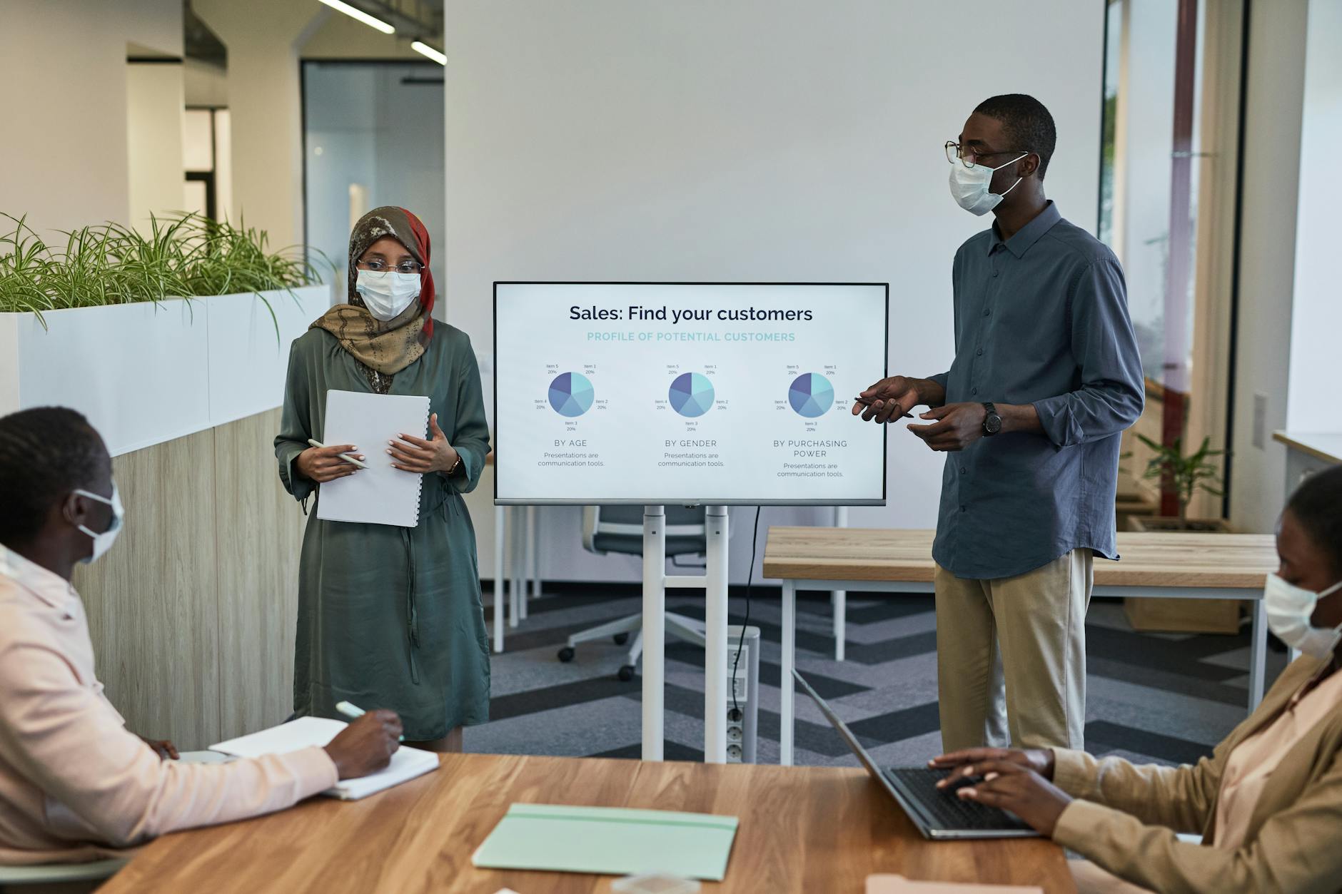 A diverse team wearing face masks discusses sales strategy with charts in a modern office.