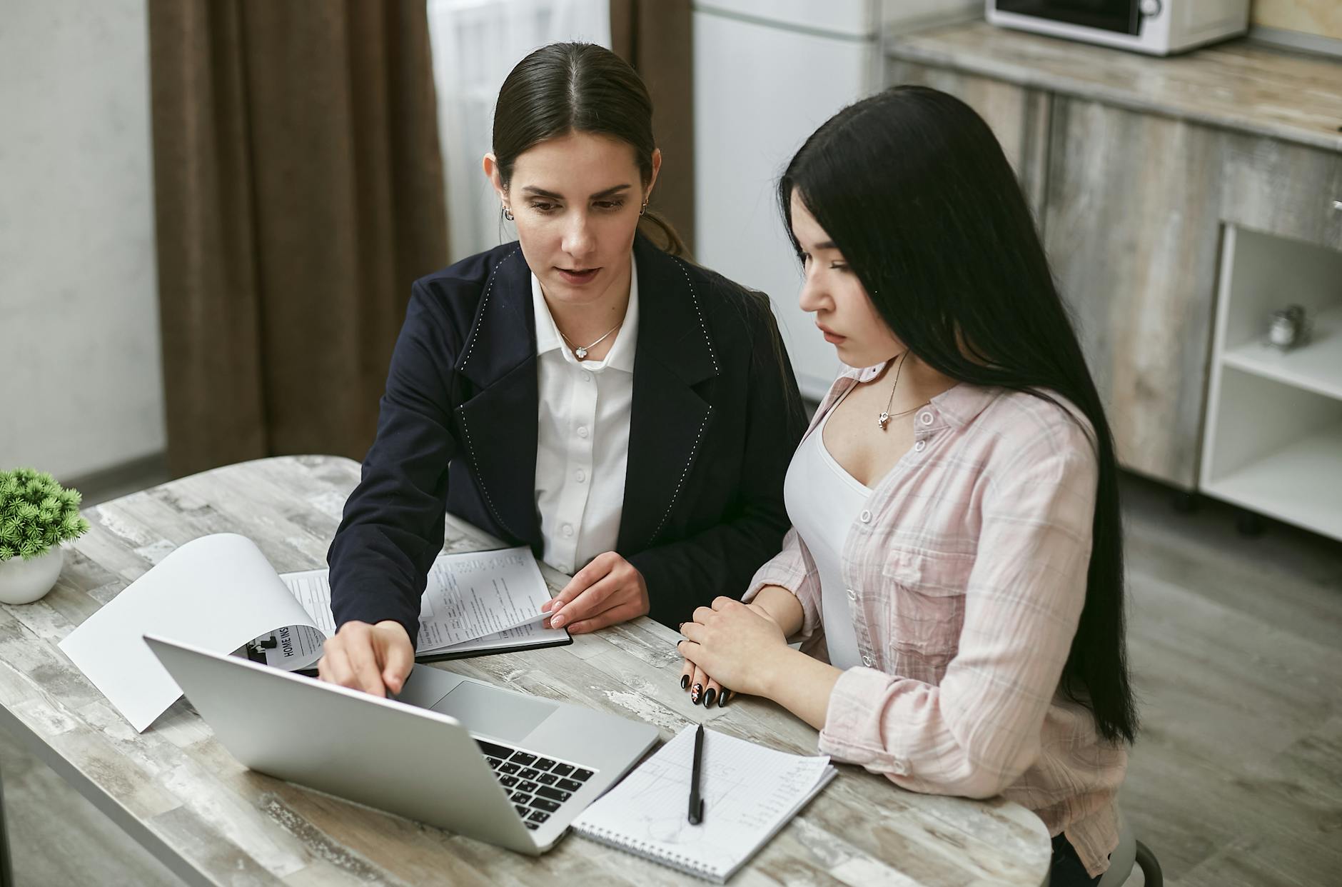 Two women working together in a modern office, focusing on a laptop and documents.