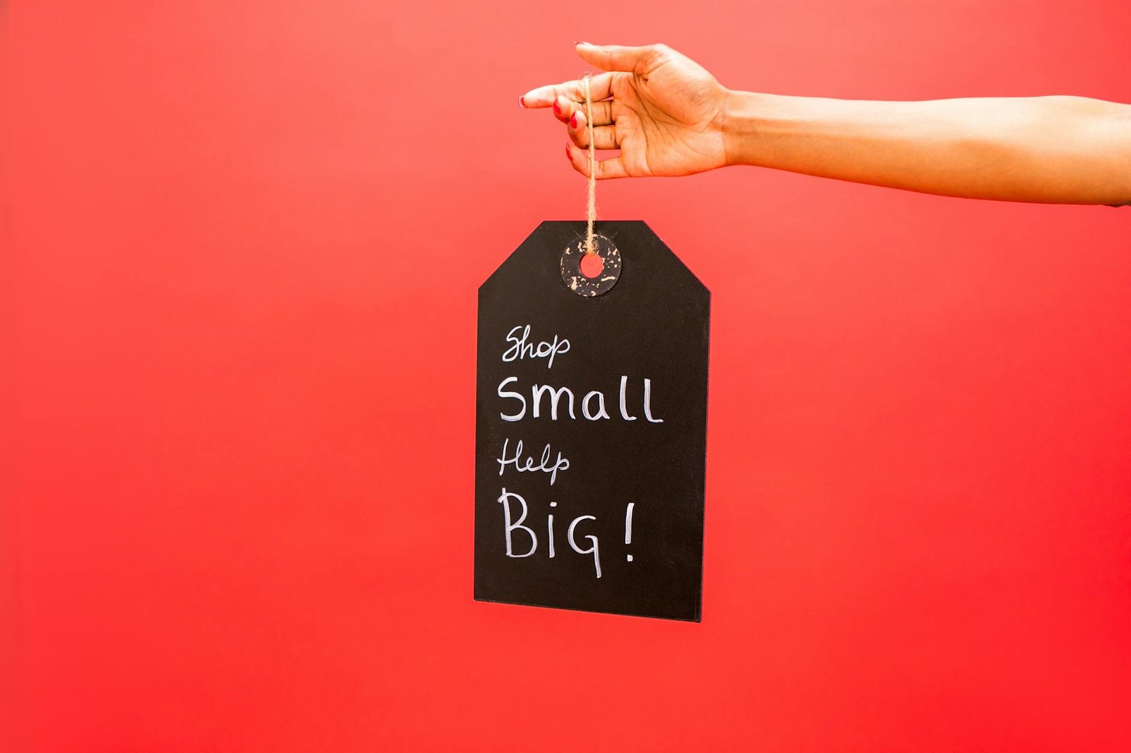 Close-up of hand holding a 'Shop Small Help Big' tag against a red background, promoting small businesses.