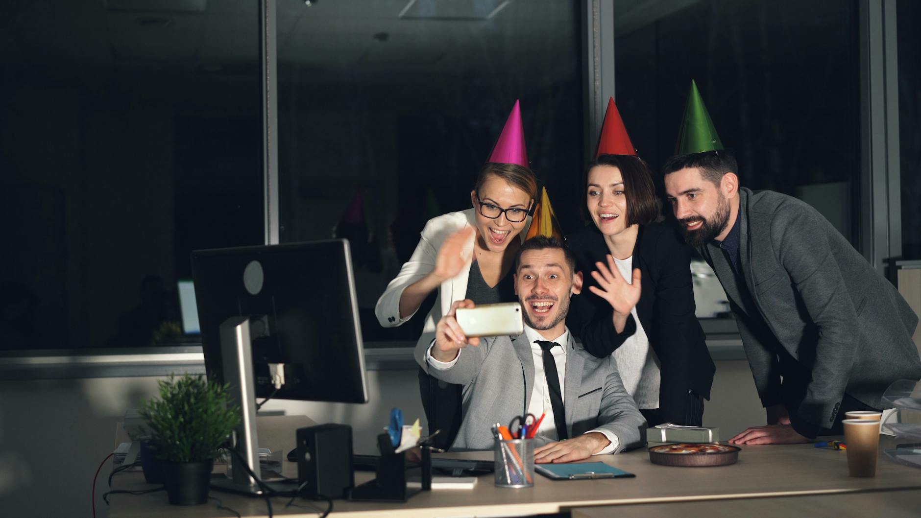 Coworkers celebrate in the office with party hats, capturing a joyful selfie.